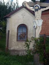 Uprosted obrzku stoj vklenkov kaplika ve patnm technickm stavu, ped n se ty krucifix s Jeem Kristem a npisem INRI. Ped kaplikou se nachz drtn plot. Vzadu je vidt zahrada se zelenm trvnkem a st rozestavnho domu.
